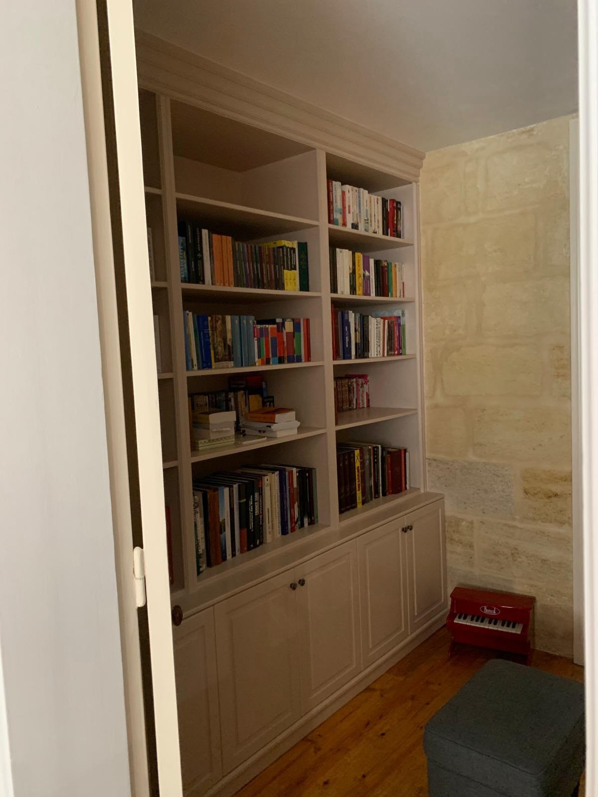 Bibliothèque sur mesure peinte gris taupe, placards moulurés en partie basse, face à un mur de pierre apparente