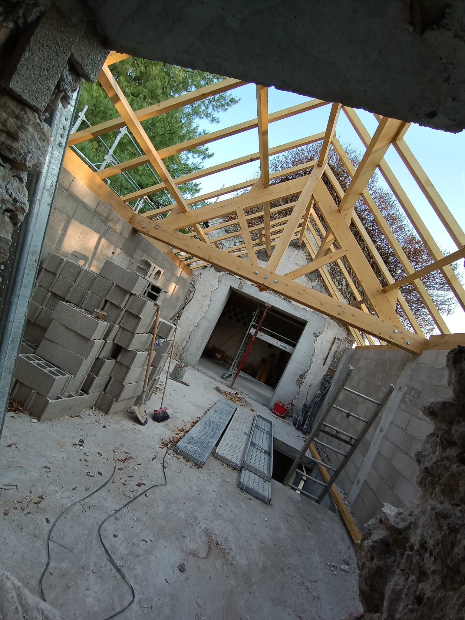 Extension de maison vue depuis une ouverture percée dans un mur ancien en pierre : charpente neuve à chevrons posée sur des murs en blocs de béton, dalle et blocs stockés au sol