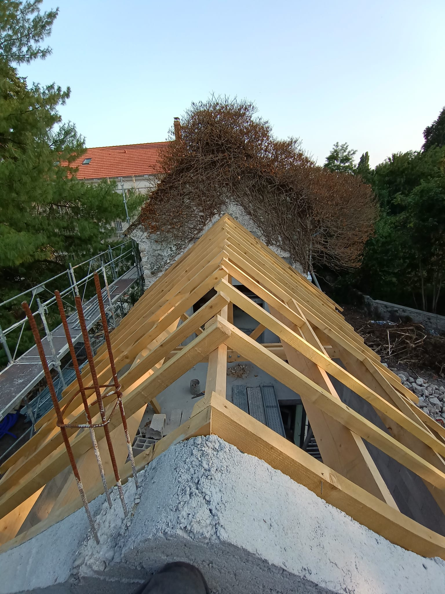 Faîtage et arêtiers d'une charpente bois neuve d'extension vus d'en haut, posés sur une tête de mur en béton aux armatures en attente, mur ancien et échafaudage en arrière-plan