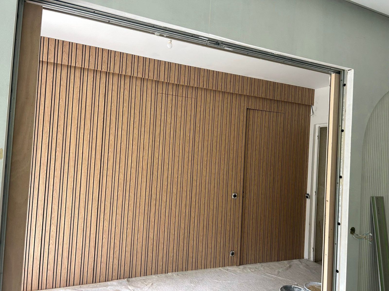 Grand panneau coulissant habillé de tasseaux en décor bois, posé sous son rail dans une ouverture à huisserie métallique, cloisons voisines encore en plaques hydrofuges vertes