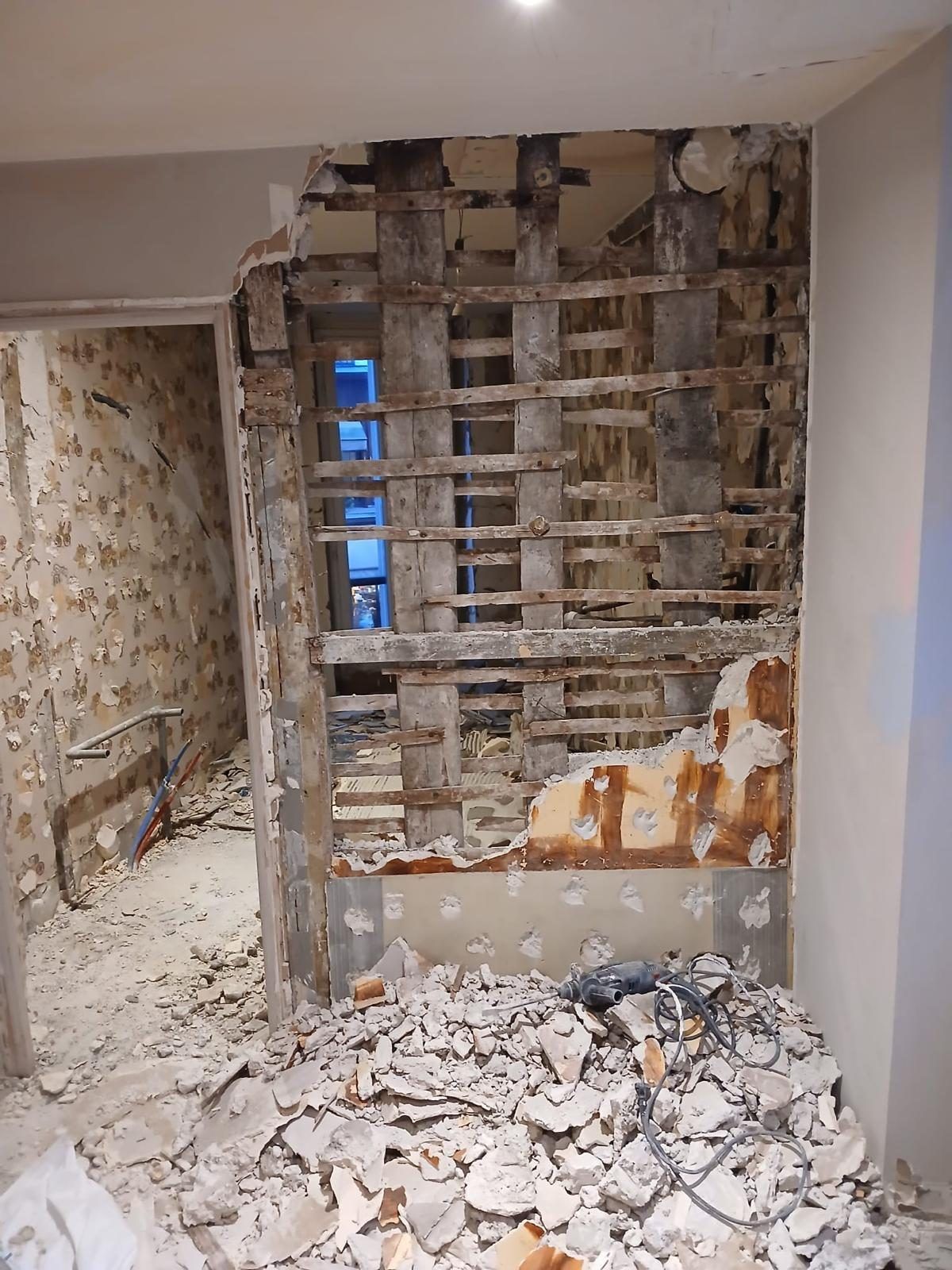 Cloison ouverte jusqu'à son ossature : lattis et bois de structure apparaissent sous le plâtre tombé au sol. C'est seulement à ce moment-là que la nature réelle du mur se lit. Chantier réel des équipes partenaires.