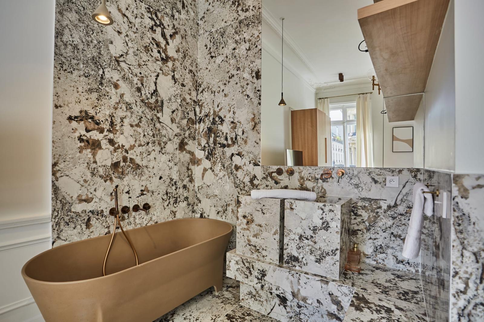 Salle de bain réelle en pierre claire à fond blanc et inclusions grises et brunes, posée du sol aux murs, avec vasque en dalles assemblées et baignoire îlot