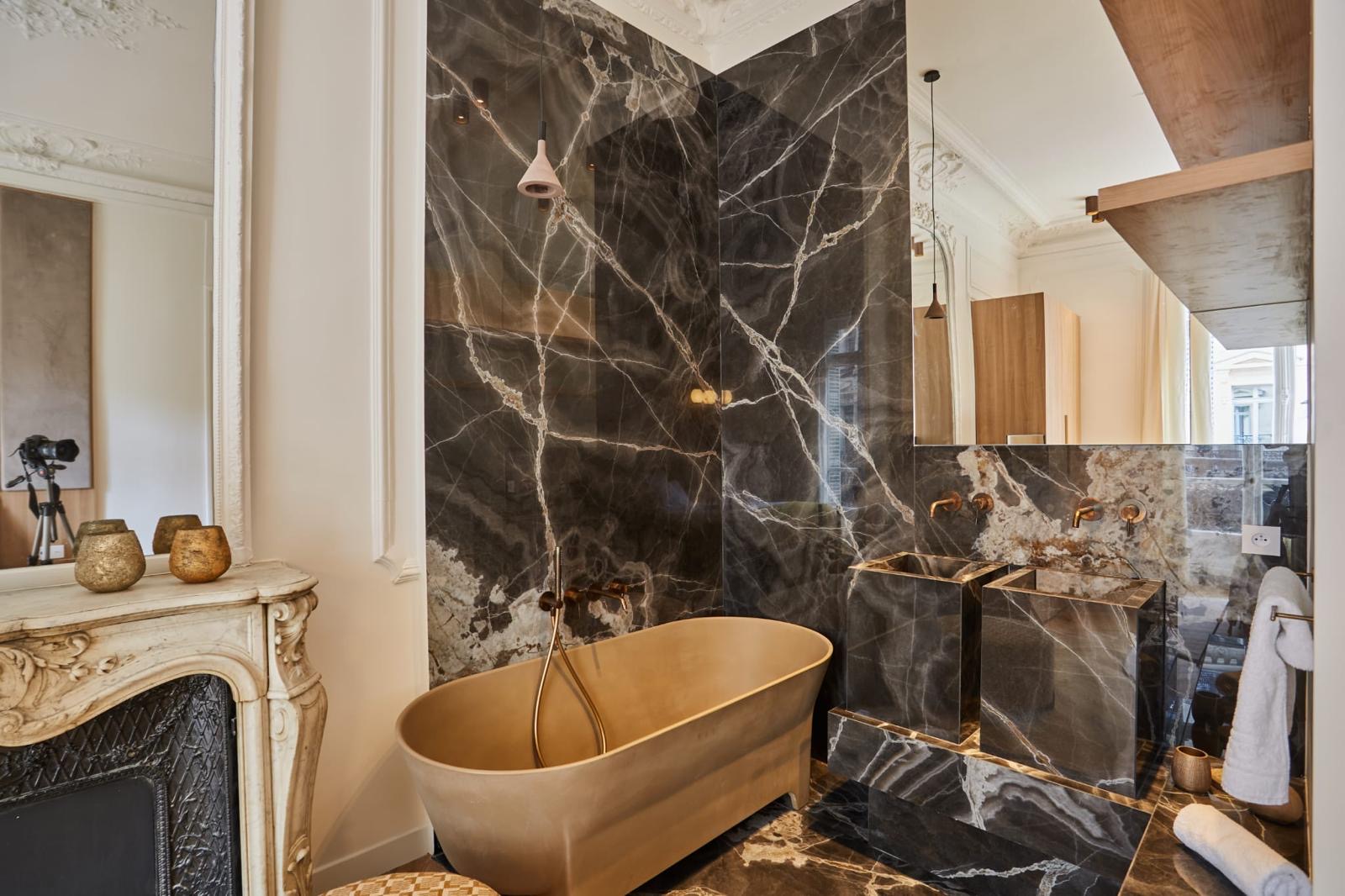 Gamme haut de gamme : marbre sombre veiné du sol au plafond, baignoire îlot et robinetterie en laiton, double vasque monolithe, cheminée de marbre d'origine conservée. Chantier réel des équipes partenaires.