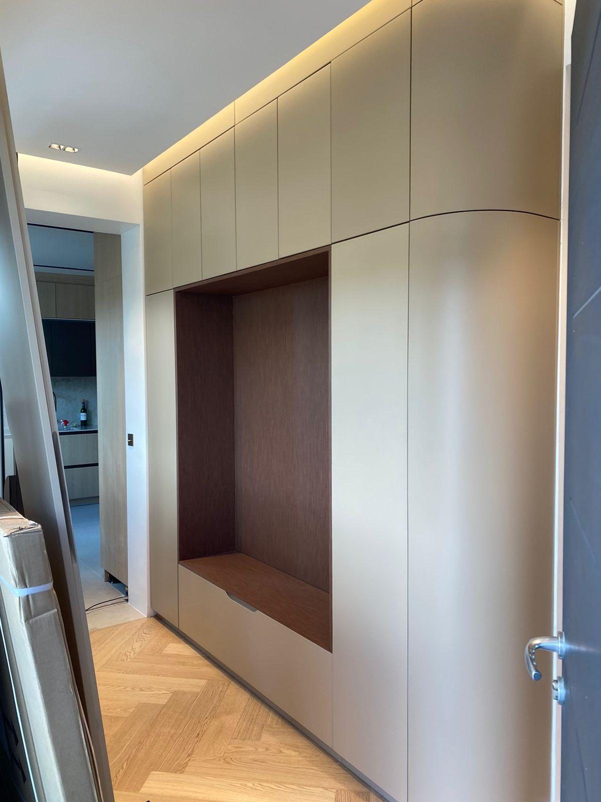 Couloir bordé d'une menuiserie toute hauteur beige à angle arrondi : un bandeau lumineux dissimulé au-dessus des portes éclaire le plafond, niche habillée de bois avec assise et tiroir