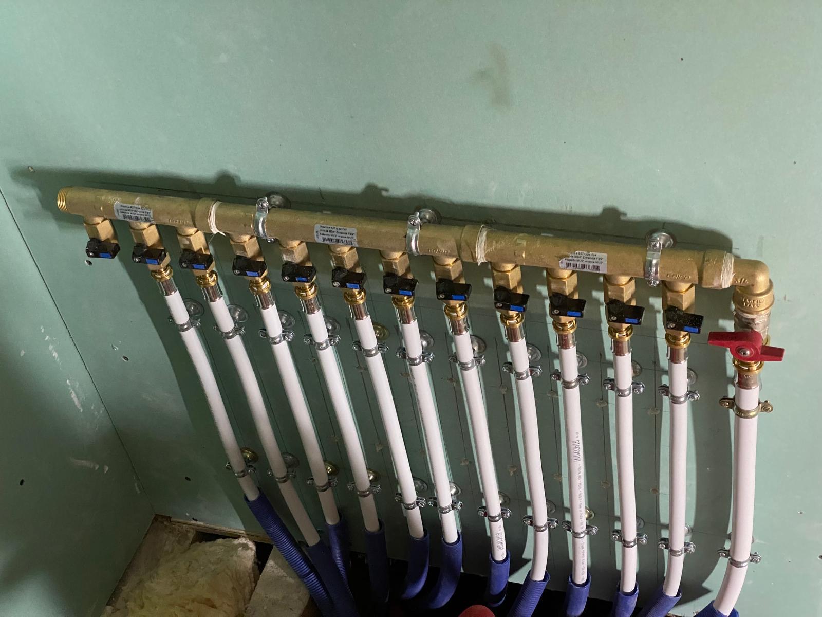 Nourrice de distribution en laiton fixée sur une plaque hydrofuge : chaque départ porte son étiquette et sa propre vanne d'isolement, la rouge commandant l'ensemble. C'est l'image à prendre avant que le doublage ne referme le mur. Chantier réel des équipes partenaires.