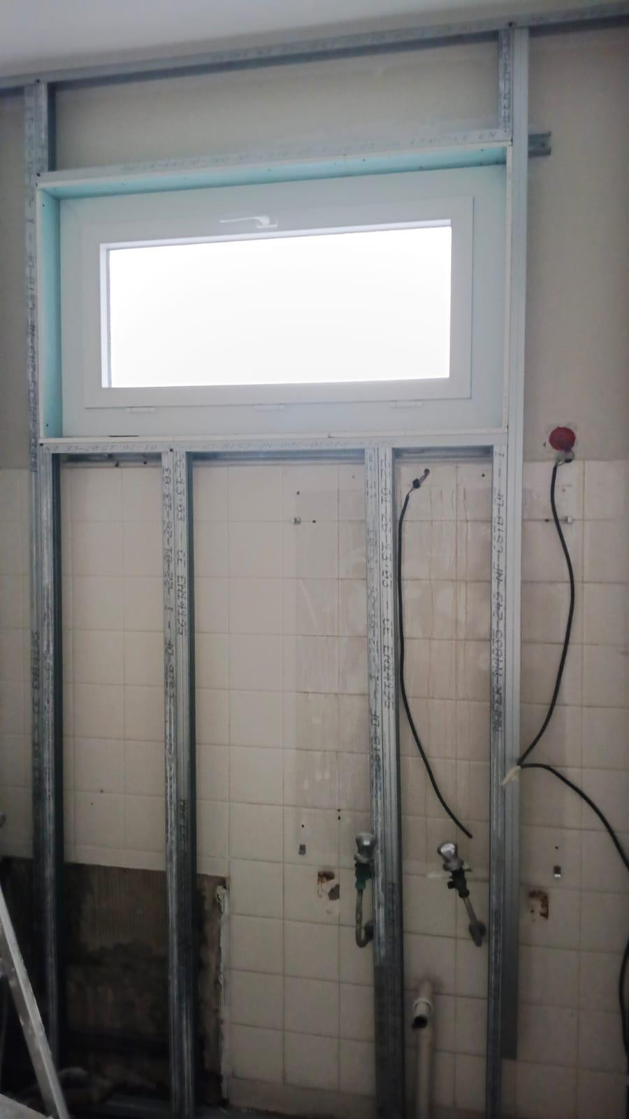 Ossature métallique montée devant un mur d'origine carrelé de faïence blanche, robinets d'arrêt, attente d'évacuation en PVC et boîte électrique encore apparents entre les montants