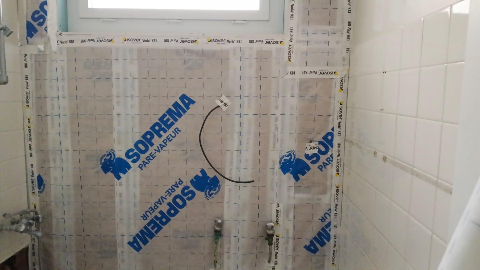 Mur d'une salle d'eau en rénovation recouvert d'une membrane pare-vapeur agrafée et adhésivée, alimentations d'eau en attente, carrelage blanc d'origine conservé à droite