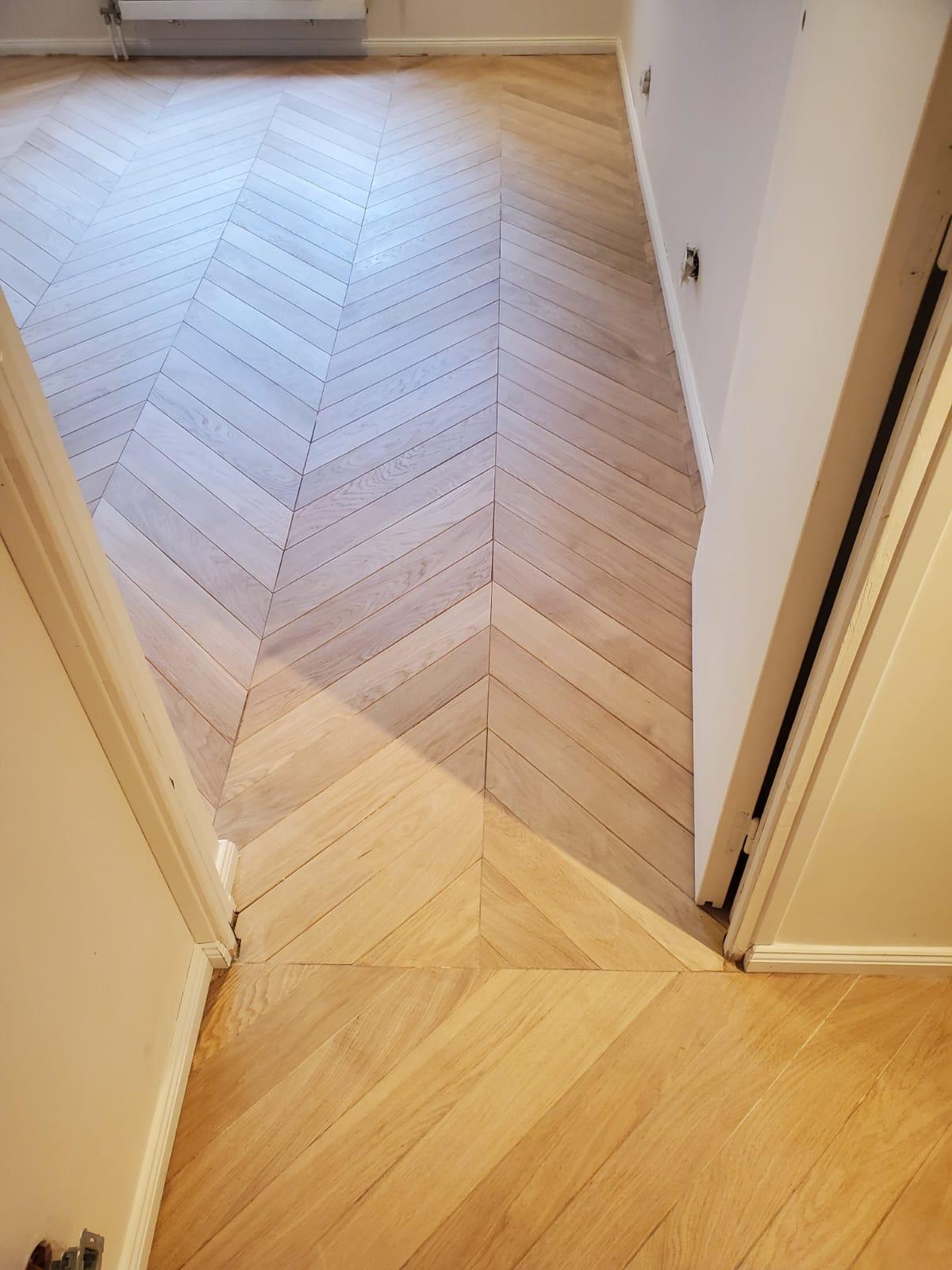 Vue rapprochée d'un parquet chêne en point de Hongrie dans un passage de porte : lames coupées d'onglet réunies par une seule ligne continue, et raccord entre deux teintes de finition