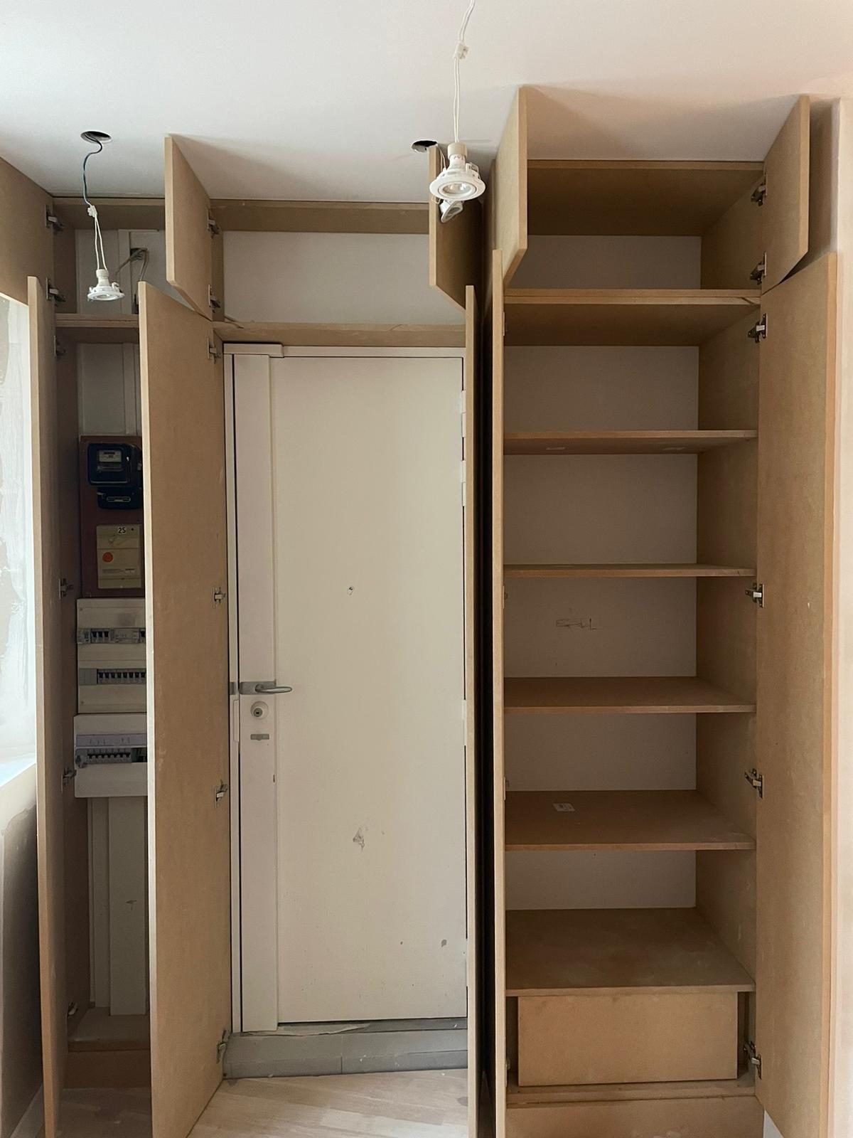 Placard d'entrée en MDF brut, portes ouvertes : une colonne abrite un compteur ancien et deux tableaux de protection, l'autre reçoit des étagères