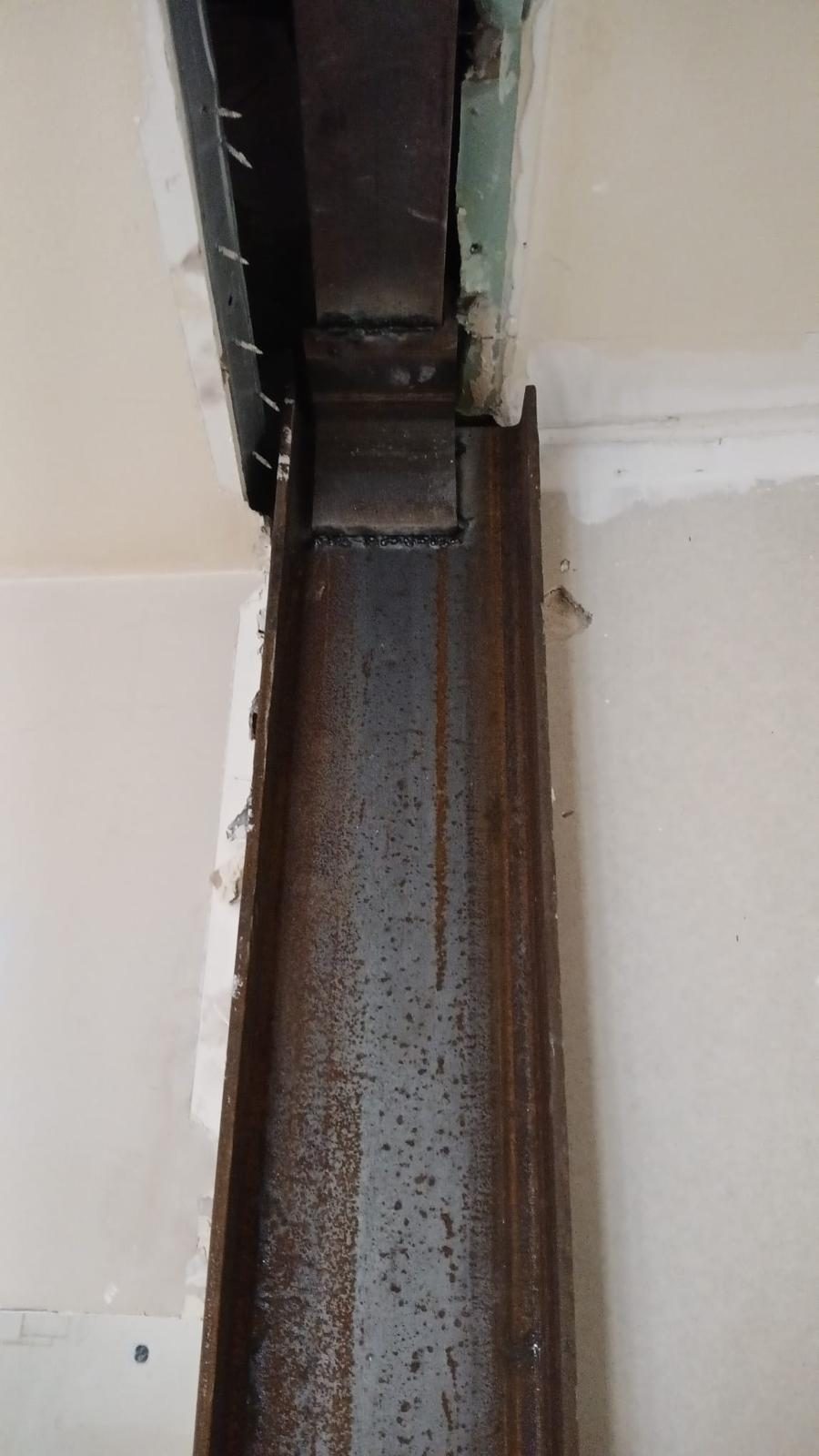 Gros plan sur la jonction soudée entre une poutre et un poteau acier, cordons de soudure visibles, plafond ouvert au-dessus