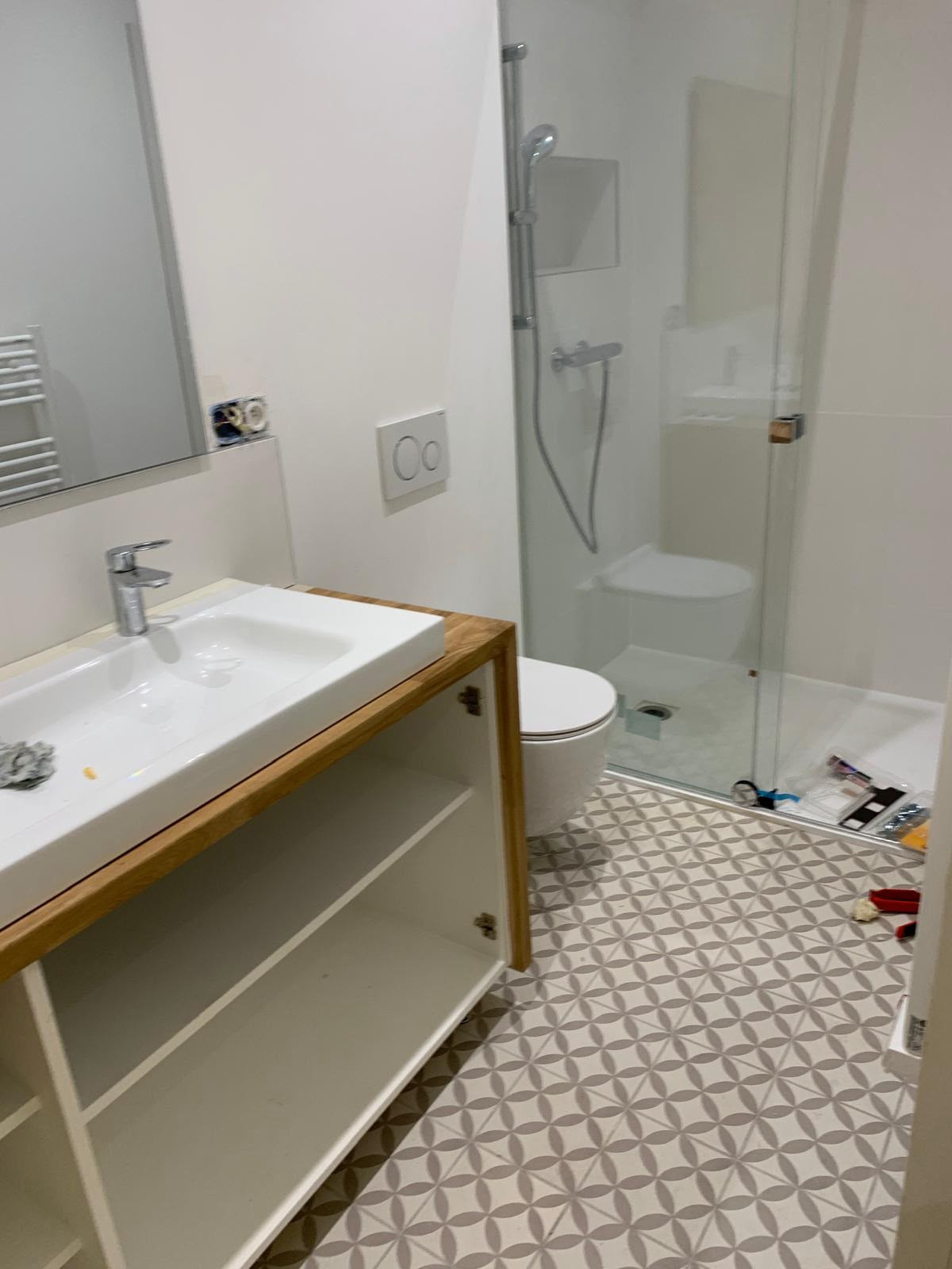 Salle d'eau d'un appartement locatif en fin de rénovation : douche à receveur extra-plat et paroi vitrée, WC suspendu, meuble vasque en bois, carreaux de ciment gris clair au sol