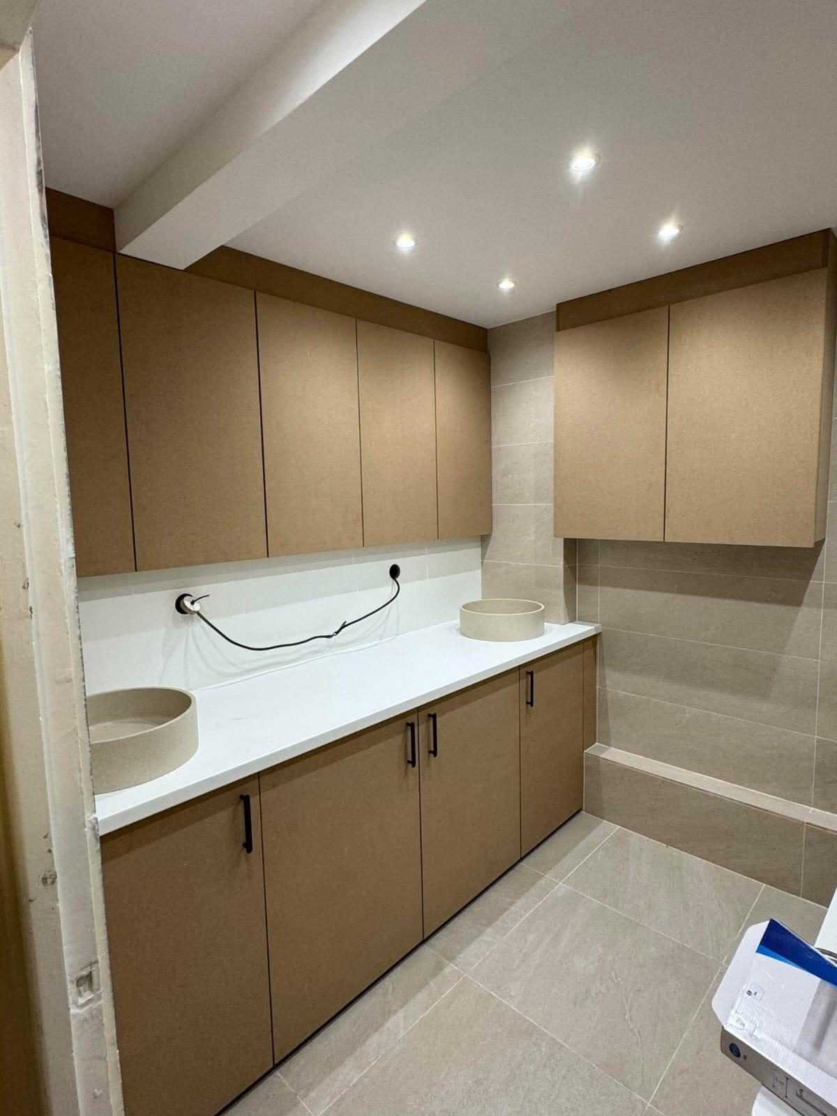 Salle d'eau en cours de finition : meubles hauts et bas en MDF brut, plan blanc et deux vasques rondes posées, faïence et sol en grand format beige, câble électrique encore pendant