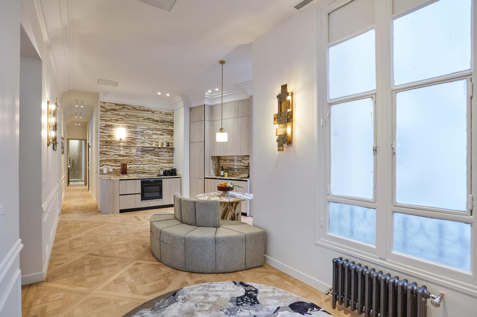 Pièce de vie haussmannienne en enfilade : cuisine en bois clair avec four encastré et crédence en pierre rubanée, banquette arrondie, parquet de Versailles et radiateur en fonte