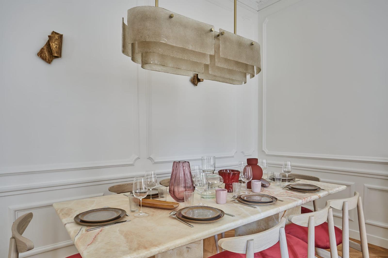 Salle à manger d'un appartement rénové tous corps d'état : table en onyx sous une suspension en verre, murs à moulures repeints en blanc, parquet chêne à chevrons