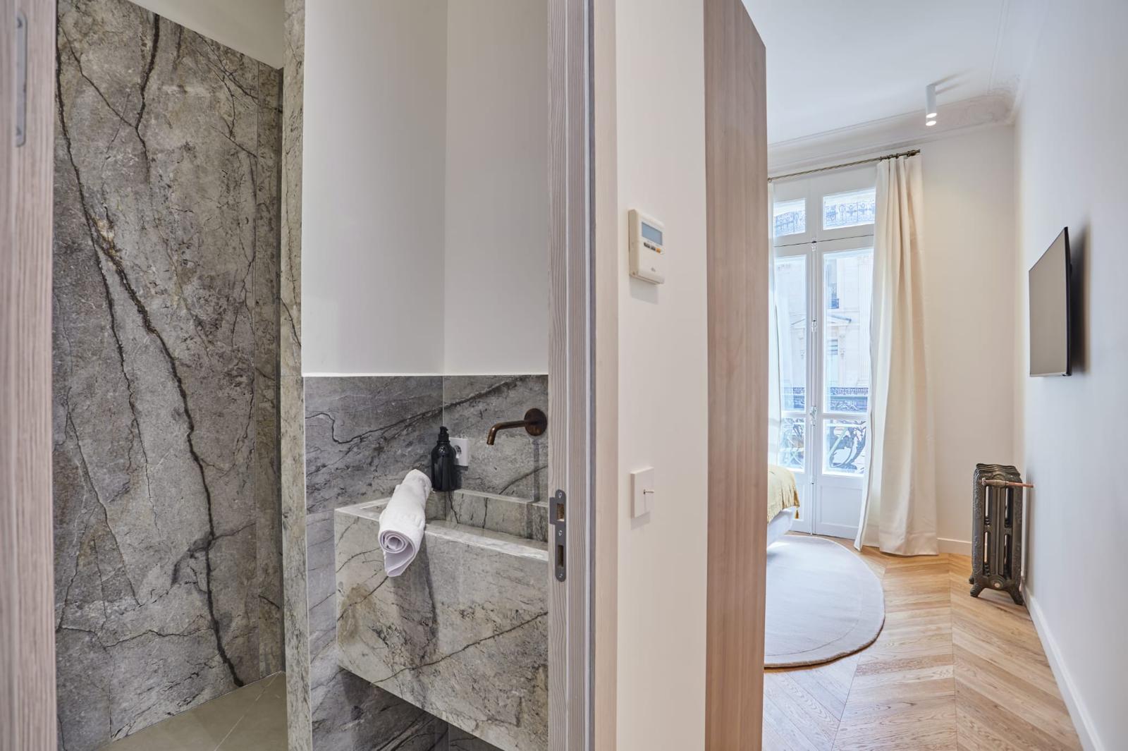 Salle d'eau en carrelage effet marbre gris veiné, vasque monolithe et robinet mural cuivré, porte ouverte sur une chambre haussmannienne au parquet à chevrons