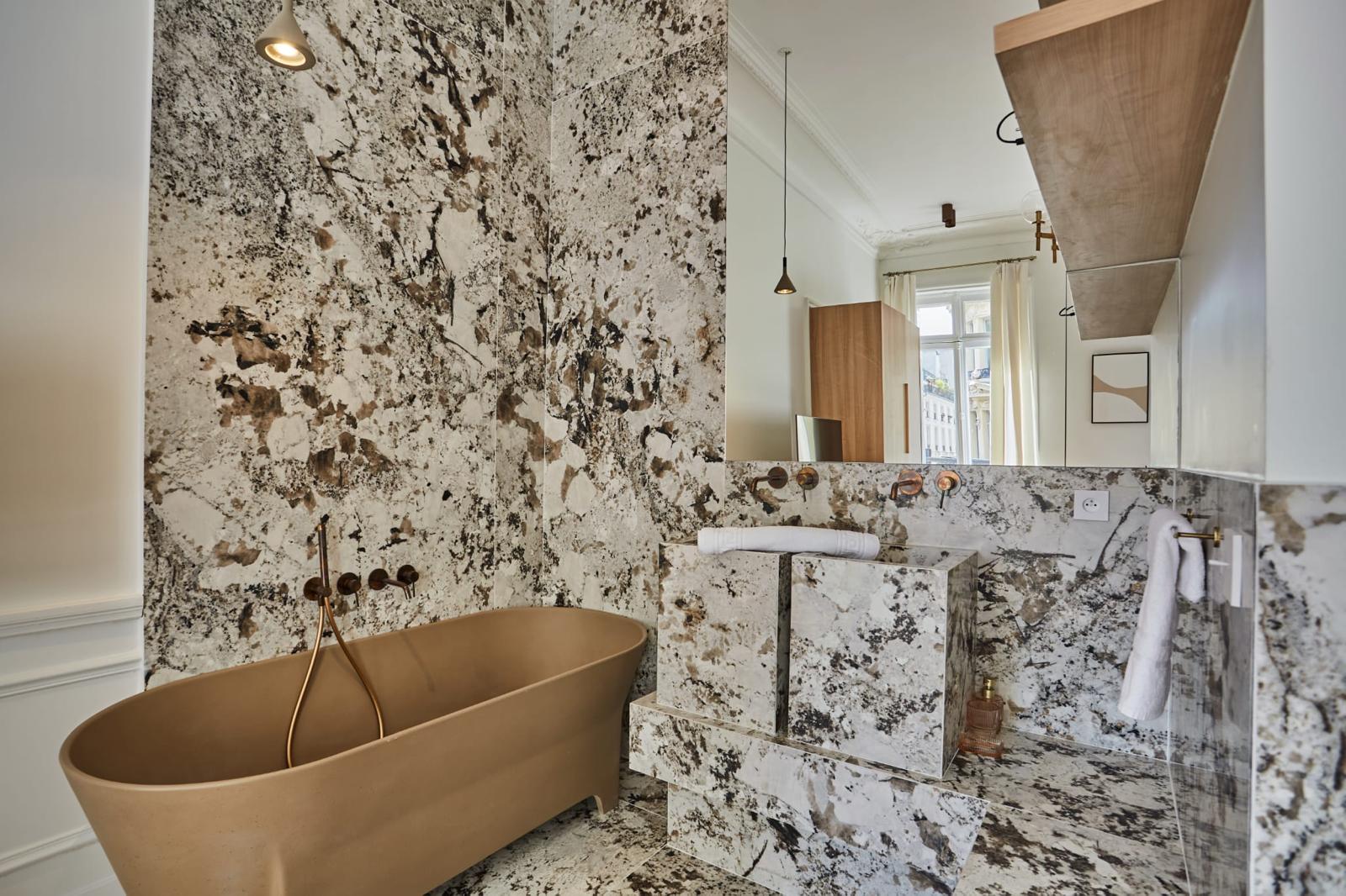 Salle de bain rénovée en granit clair avec baignoire îlot et robinetterie murale cuivrée