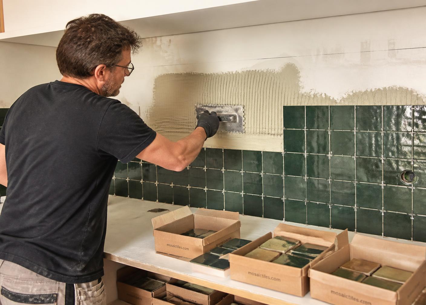Carreleur encollant au peigne le haut d'une crédence en carreaux émaillés vert foncé, croisillons de calage en place et boîtes de carreaux ouvertes sur le plan de travail