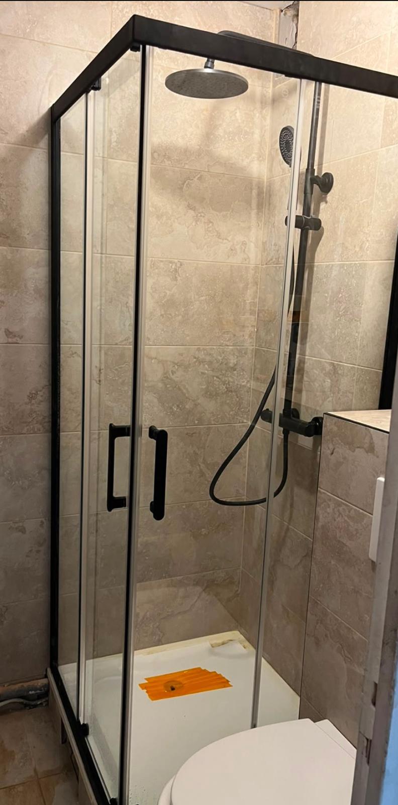Cabine de douche d'angle à parois coulissantes et profilés noirs, murs carrelés en grand format effet pierre beige, receveur blanc dont la bonde est encore protégée
